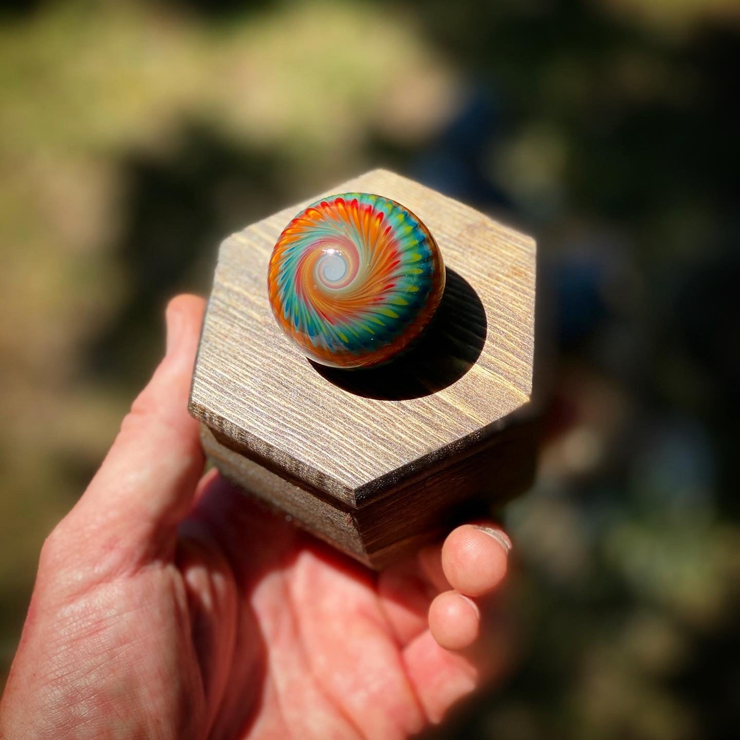 A hand is shown in the sun holding a hexagonal wooden display with a tie dye marble sitting on top. 
