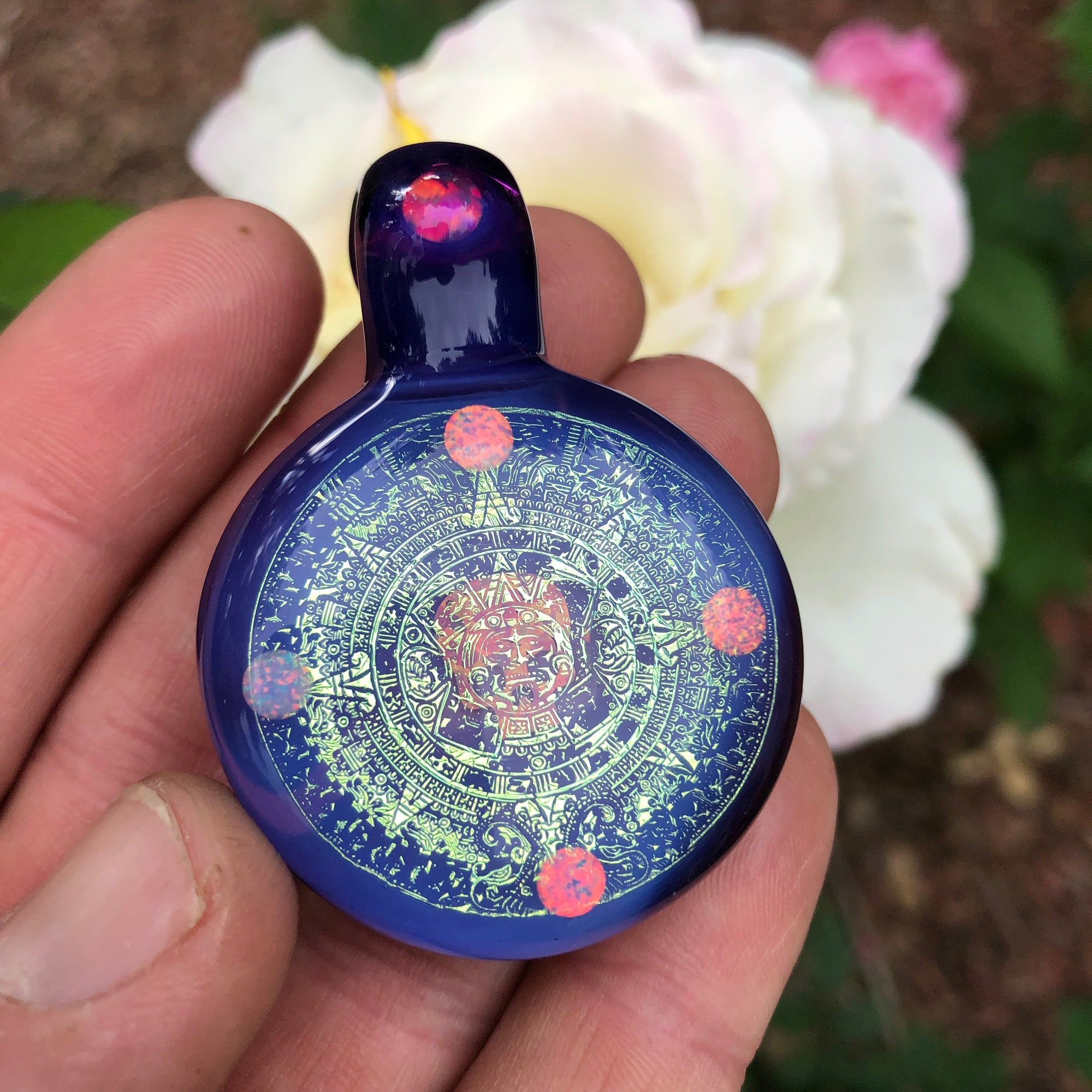 A purple dichroic hollagram pendant is held outside in front of a white flower, the pendant has  six inlaid opals, four around the face of the pendant and one on the bail, there is also a skull opal in the center of the pendant face.