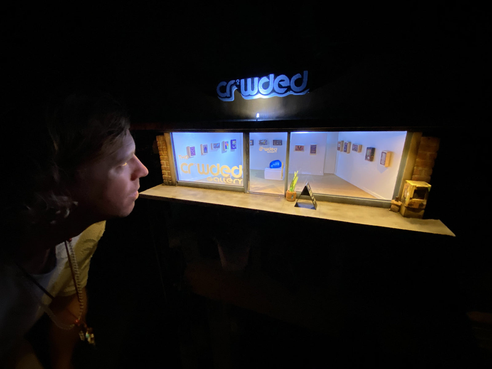 R3G15 looking into the crowded gallery from the front left side of the model, the area is dark except for the lights inside the miniature model that light up R3G15 face. 