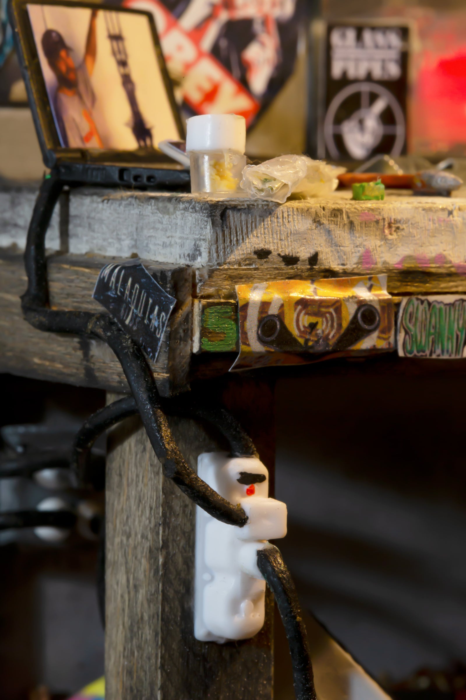 Close up photo of the miniature glassblowing station, showing the bench and a laptop, there is a bag of herb on the bench as well. 
