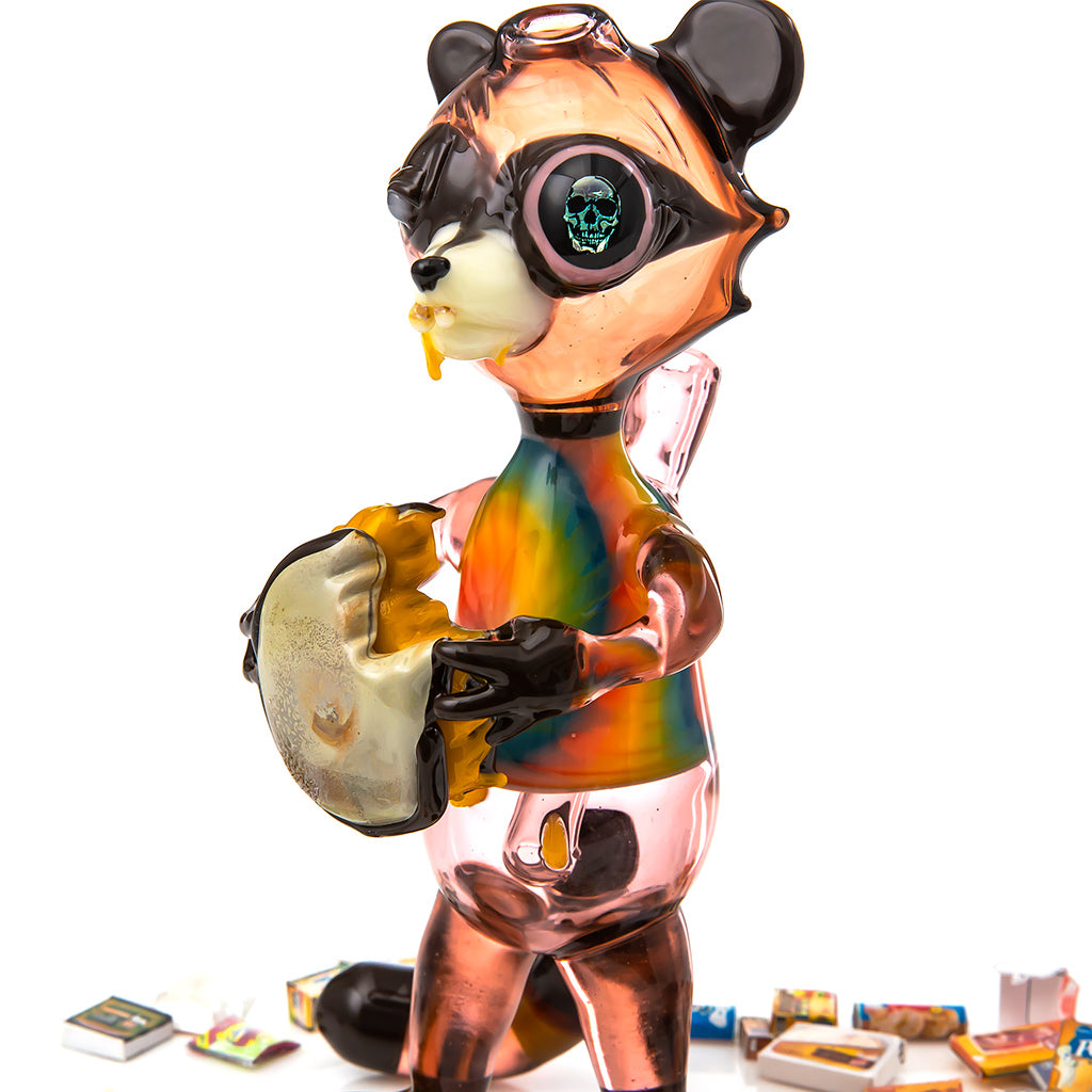 Close up studio photo showing a heady glass piece by R3G15, a Raccoon wearing a tie dye shirt is eating a grilled cheese sandwich, he has a dichroic skull image as an eye patch. 