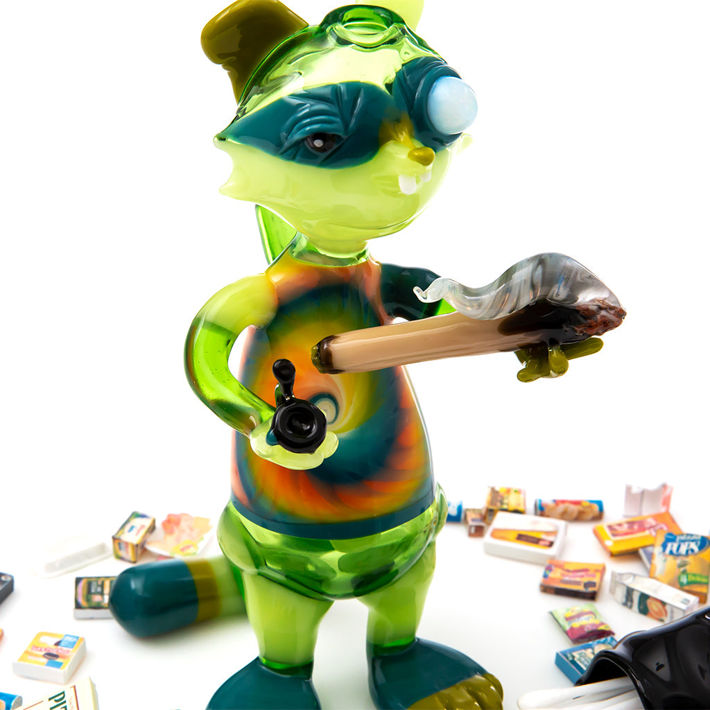 An incredible glass blown sculpture of a raccoon smoking a joint in one hand and a pipe in the other. The raccoon is bright green with an opal in his left eye, he has a tie dye shirt and is standing among trash.